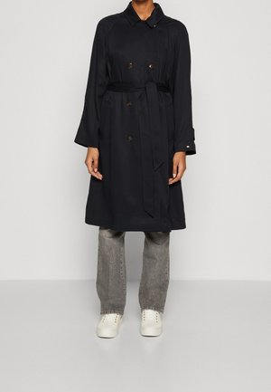 Person iført en sort dobbeltradet trenchcoat med bælte, grå jeans og hvide sneakers, der står mod en ensfarvet grå baggrund.