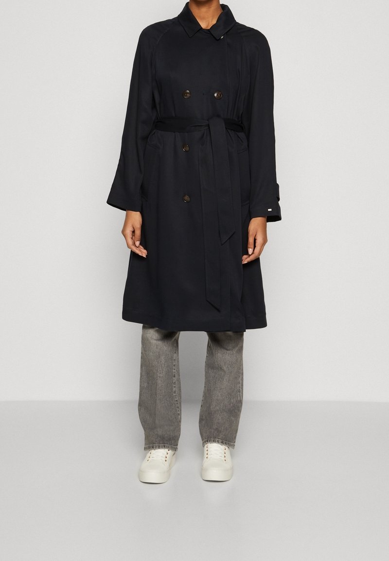 Personne portant un trench-coat noir croisé avec ceinture, un jean gris et des baskets blanches, debout devant un fond gris uni.