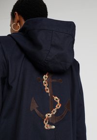 Person med kort lockigt hår bär en mörk huva jacka som har en stor broderad ankare- och kedjedesign på ryggen.