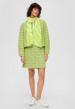 Grüne strukturierte Jacke und passende Rock mit einem weißen Akzent-Schal. Das Outfit hat einen lässigen Schnitt und einfache weiße Sneaker.