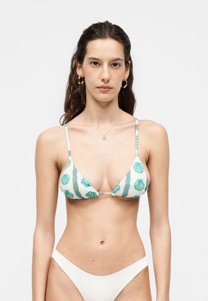 Tânără cu păr lung și închis la culoare, purtând cercei cerc aurii, un colier, un top de bikini alb cu imprimeuri albastre cu scoici și slip alb de bikini asortat.