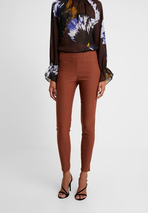 Leggings marron extensibles avec une coupe ajustée, dotés d'une taille élastique. Associés à un chemisier transparent à manches longues et à motifs. Talons noirs.