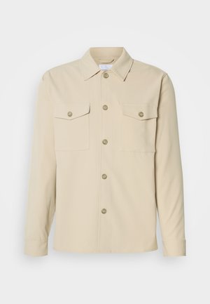 Beige overhemd met knopen, gemaakt van een gladde stof, heeft twee borstzakken, een klassieke kraag en vijf knopen aan de voorkant. Lange mouwen met knoopmanchetten.