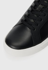 Zwarte leren sneaker met een ronde neus, voorzien van platte zwarte veters, een gladde oppervlakte en een witte rubberen zool met textuurafwerking.