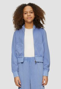 Blaue, kurz geschnittene Zip-Jacke aus weichem Material mit zwei vorderen Taschen, gerippten Bündchen und einem hohen Kragen. Kombiniert mit passenden blauen Hosen.