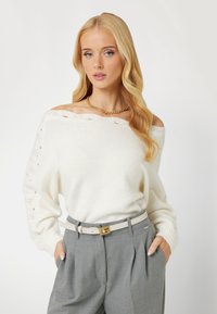 Maglione bianco off-the-shoulder con motivo a maglia testurizzato e polsini a costine, abbinato a pantaloni grigi a vita alta e una cintura leggera.