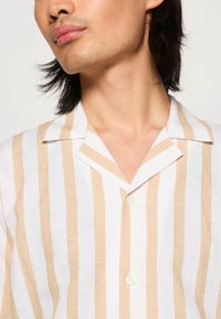 Kevyt, kauluksellinen paita valkoisena, jossa on pystysuoria beigejä raitoja. Varustettu nappilista ja pehmeä, teksturoitu kangas.