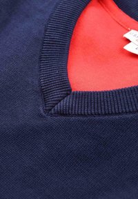 Nahaufnahme eines marineblauen Strickpullovers mit einem gerippten V-Ausschnitt, der ein rotes Innenhemd und ein kleines weißes Kleidungsetikett zeigt.