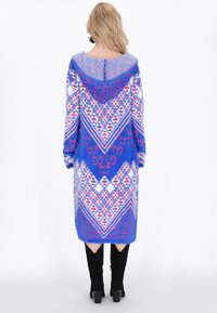 Long cardigan à capuche en tissu bleu moelleux avec des motifs géométriques blancs et rouges, offrant une coupe décontractée et une longueur mi-mollet.