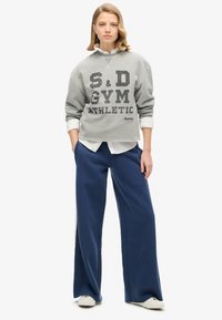 Grijze sweatshirt met vetgedrukt zwart tekst "S&D GYM ATHLETIC" en een wit overhemd eronder; gecombineerd met losse blauwe joggingsbroek en witte schoenen.