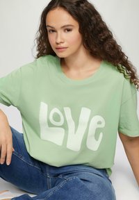 Jasnozielona bawełniana koszulka z dużym białym napisem „LOVE”. Krótkie rękawy, okrągły dekolt, luźny krój. Model z pozą siedzącą w dżinsach.