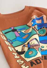 Bruin shirt met kleurrijke cartoonillustraties van een blauwe helikopter en een blauwe tractor met tekst "ENGINE RISE" en gedeeltelijk zichtbare letters.