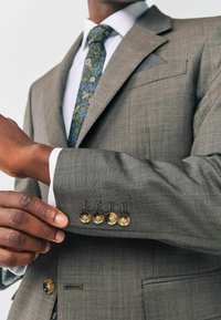 Grau maßgeschneiderte Sakkojacke aus strukturiertem Stoff, mit Reverskragen und vier goldenen Knöpfen an den Ärmeln, darunter eine Krawatte mit Blumenmuster.