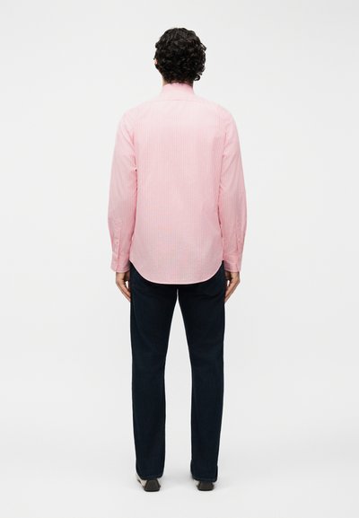 Άνδρας με σγουρά μαλλιά, φορούσε ροζ ριγέ πουκάμισο με μακριά μανίκια και σκούρο παντελόνι, στέκεται γυρισμένος πλάτη σε λευκό φόντο.