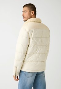 Giubbotto imbottito color crema chiaro con colletto in peluche. Presenta una texture trapuntata, polsini con zip e una vestibilità comoda.