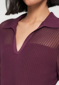 Vinrød strikket topp med teksturert ribbestrikket design, har V-hals og gjennomsiktige stripete ermer. Mykt stoff med en litt tettsittende silhuett.