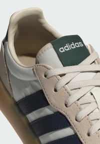 Bílá a béžová teniska se zelenými akcenty, s třemi tmavě modrými pruhy a bílými tkaničkami. Kvalitní kožený svršek a strukturovaná podrážka.