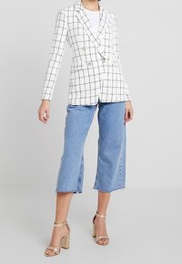 White double-breasted blazer with black check pattern, paired with cropped light blue denim pants, and gold block-heel sandals.