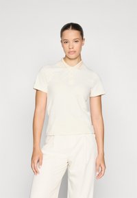 Krem polo skjorte med krage og tre knapper foran. Har et brodert logo i lys oransje. Lett stoff og korte ermer.