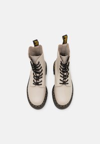Botas de tobillo de cuero beige con cordones negros, puntera redonda, acentos de costura amarilla y una lengüeta amarilla en el talón. Forro de lona visible en el interior.