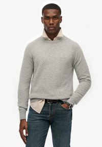 Helles graues Strickpullover mit hohem Kragen, getragen über einem beigefarbenen Hemd, kombiniert mit dunkelblauen Jeans und einem schwarzen Gürtel.