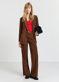 Brauner Cordblazer mit Doppelknopfreihe, kombiniert mit passenden hoch taillierten Hosen. Ein rotes Oberteil und eine schwarze Handtasche vervollständigen das Outfit.