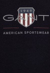 Dunkelblaues Baumwoll-T-Shirt mit einem Schild-Logo, das rot-weiße Streifen zeigt, mit dem Text "GANT" darüber und "AMERICAN SPORTSWEAR" darunter.