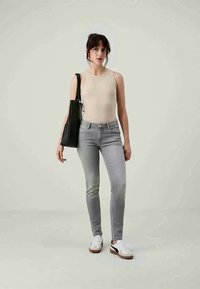 Žena v béžovém bezrukávovém topu, šedých slim-fit džínách a bílých teniskách, držící černou tašku přes rameno, stojící na jednobarevném pozadí.