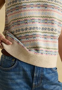 Person, der einen gemusterten Strickpullover trägt und den Saum anhebt, um blaue Jeans und einen braunen Gürtel mit einer goldenen Schnalle zu zeigen.