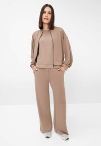 Felpa beige e pantaloni a gamba larga abbinati in tessuto morbido. La felpa presenta una chiusura con zip davanti e maniche arricciate. Un paio di sneakers bianche completano il look.