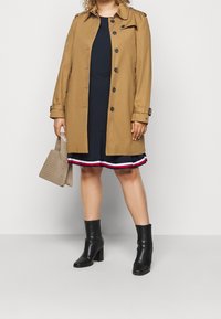Una persona está de pie con un abrigo de trench beige sobre un vestido azul marino con un dobladillo a rayas, sosteniendo un bolso texturizado y usando botas negras hasta el tobillo.