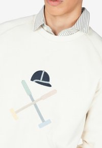 Sudadera de color claro con palas de pólo bordadas y un casco en colores pastel. Presenta un cuello redondo y un cuello a rayas debajo.