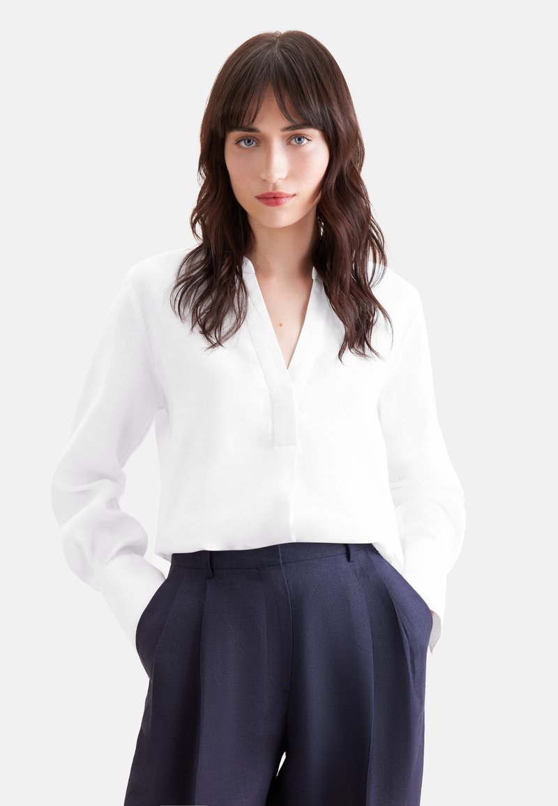 Femme aux cheveux bruns ondulés portant un chemisier blanc à manches longues et col en V, ainsi qu'un pantalon bleu marine taille haute, mains dans les poches, fond neutre.