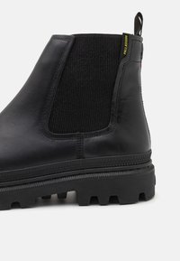 Bottine en cuir noir avec panneau latéral en élastique, semelle en caoutchouc texturé, languette de traction et accent logo contrastant en jaune et rose.