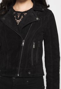 Donna che indossa una giacca da biker in suede nera con zip asimmetrica, bottoni a pressione argentati, due tasche con zip e un top in pizzo sotto.