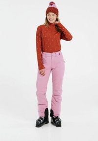 Hnedý top s dlhým rukávom a bielymi bodkami, v kombinácii s ružovými nohavicami s čiernymi akcentmi a pásom na dve gombíky. Čierne lyžiarske topánky.