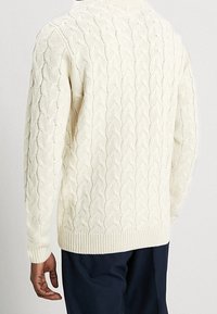 Maglione crema a maglia cable-knit con un motivo testurizzato, maniche lunghe e orlo a coste; indossato con pantaloni scuri.