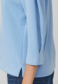 Jasnoniebieski top z rękawami do połowy przedramienia, wykonany z gładkiej tkaniny, z bocznymi rozcięciami i delikatnymi przeszyciami wzdłuż brzegu.