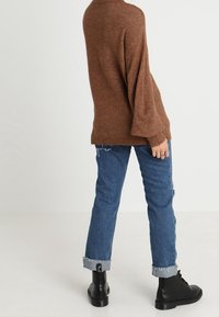 Hnědý, oversize svetr s žebrovanými manžetami, kombinovaný s trhanými, rovně střiženými modrými džíny a černými šněrovacími botami. Pohodlný, strukturovaný materiál.