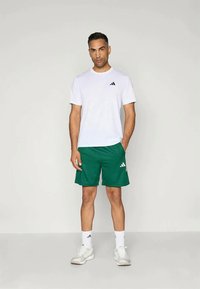 Bela kratka majica s kratkimi rokavi in črnim logotipom, v kombinaciji z zelenimi športnimi kratkimi hlačami z teksturiranim vzorcem in belimi poudarki. Bele superge.