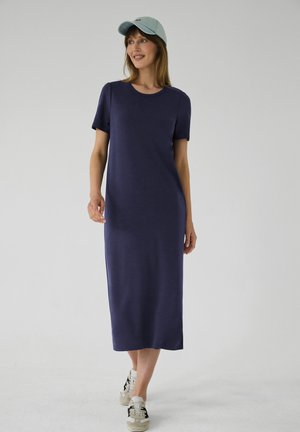 Kvinde iført en mørkeblå midi-kjole med korte ærmer, lys blå kasket og hvide sneakers, stående mod en ensfarvet lys baggrund.