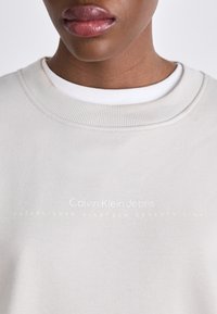 Lys beige sweatshirt med ribbet mannskapskrage. Har subtilt gulltekst: "Calvin Klein Jeans" og "ETABLERES NITTENDE ÅTTI".