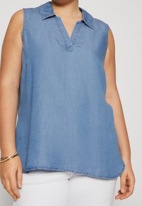 Lyseblå ærmeløs denimtop med krave, V-hals design og rundet kant. Blød tekstur med diskrete syninger.