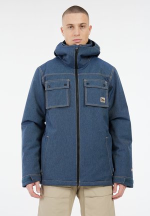 Man in een blauwe hooded jacket met een ritssluiting aan de voorkant en twee knoopzakken op de borst, gecombineerd met beige broeken, staand tegen een witte achtergrond.