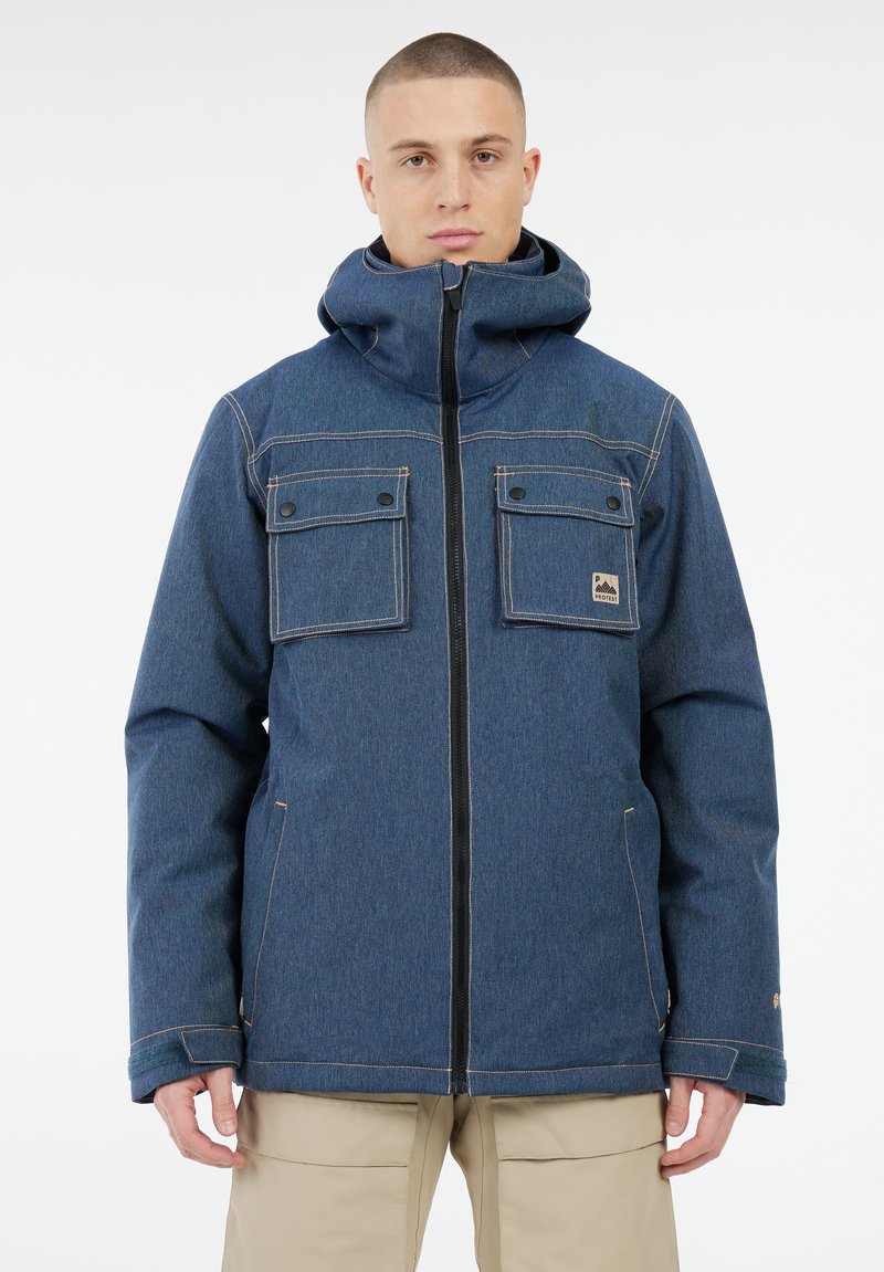 Mann in blauer Kapuzenjacke mit Reißverschluss vorne und zwei knöpfbaren Brusttaschen, kombiniert mit beigen Hosen, steht vor einem weißen Hintergrund.