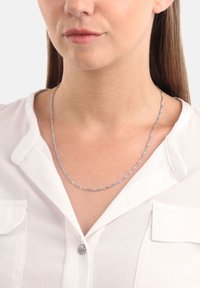 Kvinna som bär ett vridet silverkedjesmycke över en vit skjorta med knappar, med krage och bröstfickor.