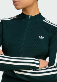 Dark green ribbed athletic jacket with white zipper and three white stripes on the sleeves. Features a small white logo on the chest.