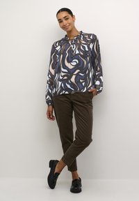 Printet bluse i mørkeblå, beige og lyseblå sværte; lange ærmer, rund hals med bindebånd; sat sammen med brune fløjl bukser og sorte sko.