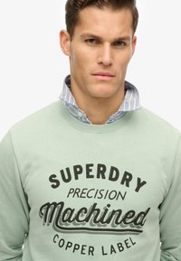 Sweatshirt vert clair avec le texte noir "Superdry Precision Machined". Porté par-dessus une chemise à col rayée bleue et blanche. Texture lisse.