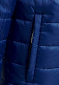 Chaqueta azul acolchada que presenta un tejido de nylon suave y texturizado con un bolsillo con cremallera. Los paneles cosidos crean una apariencia de acolchado.